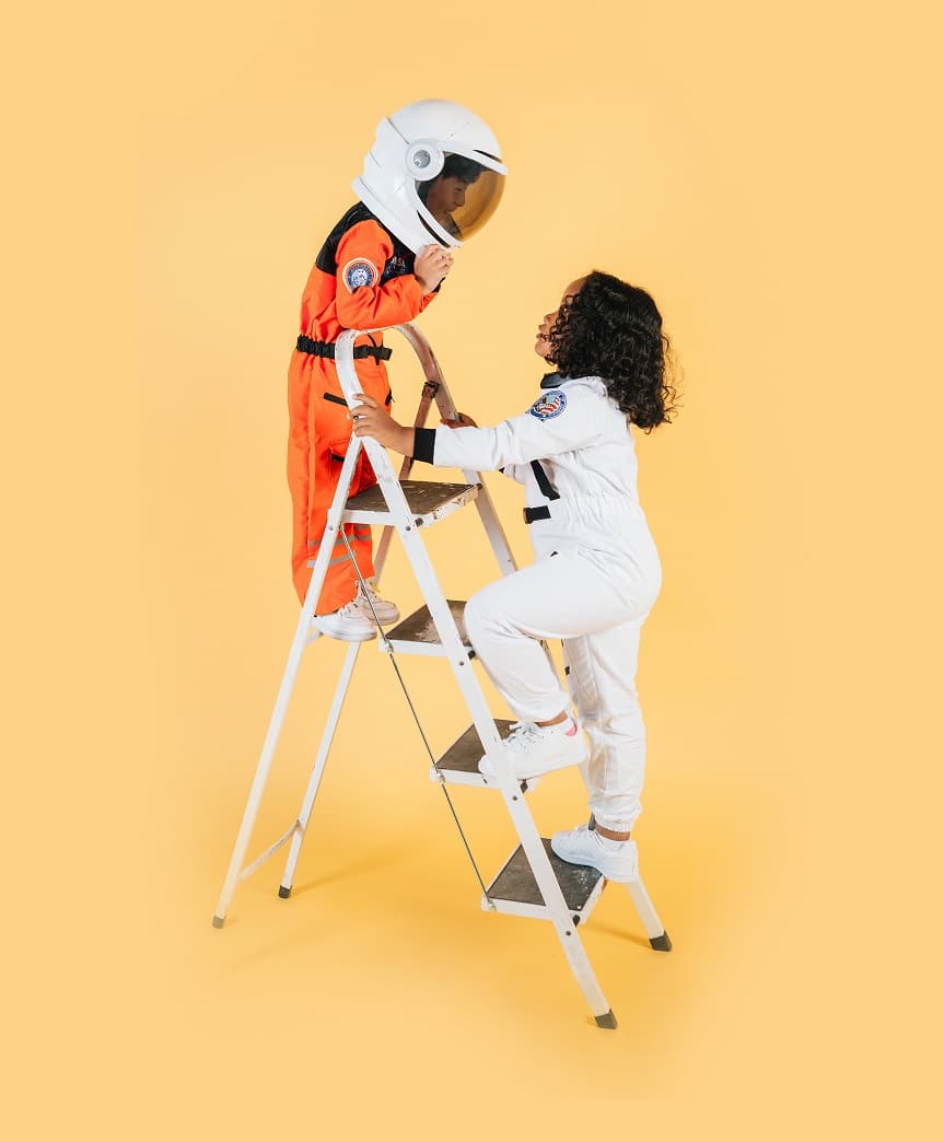 A child in an astronaut suit climbing a ladder with the assistance of another child, symbolizing teamwork, growth, and the development of responsibility through Shop On Fire’s 12X Parenting Coaching.